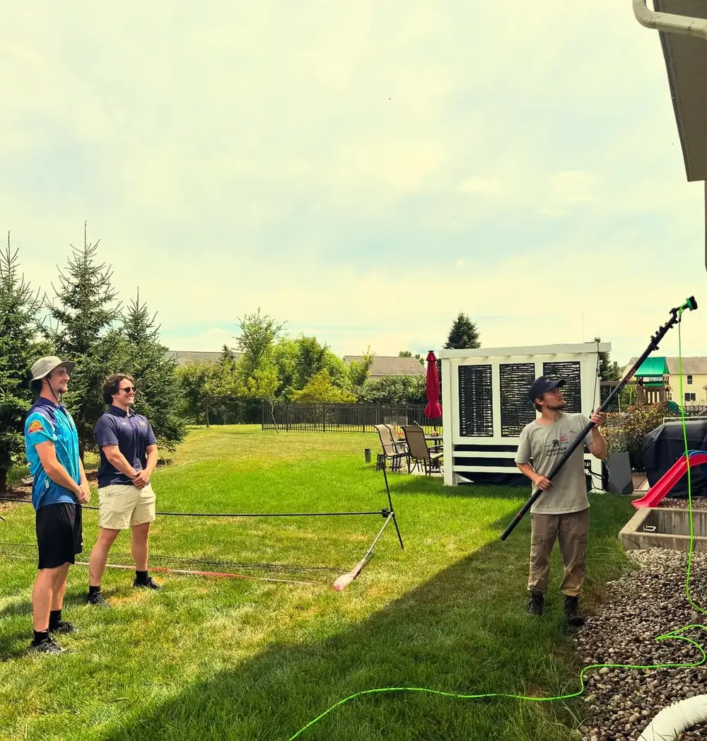 Technician rinsing a home's eaves with an extension pole while two teammates look on in Okemos, MI
