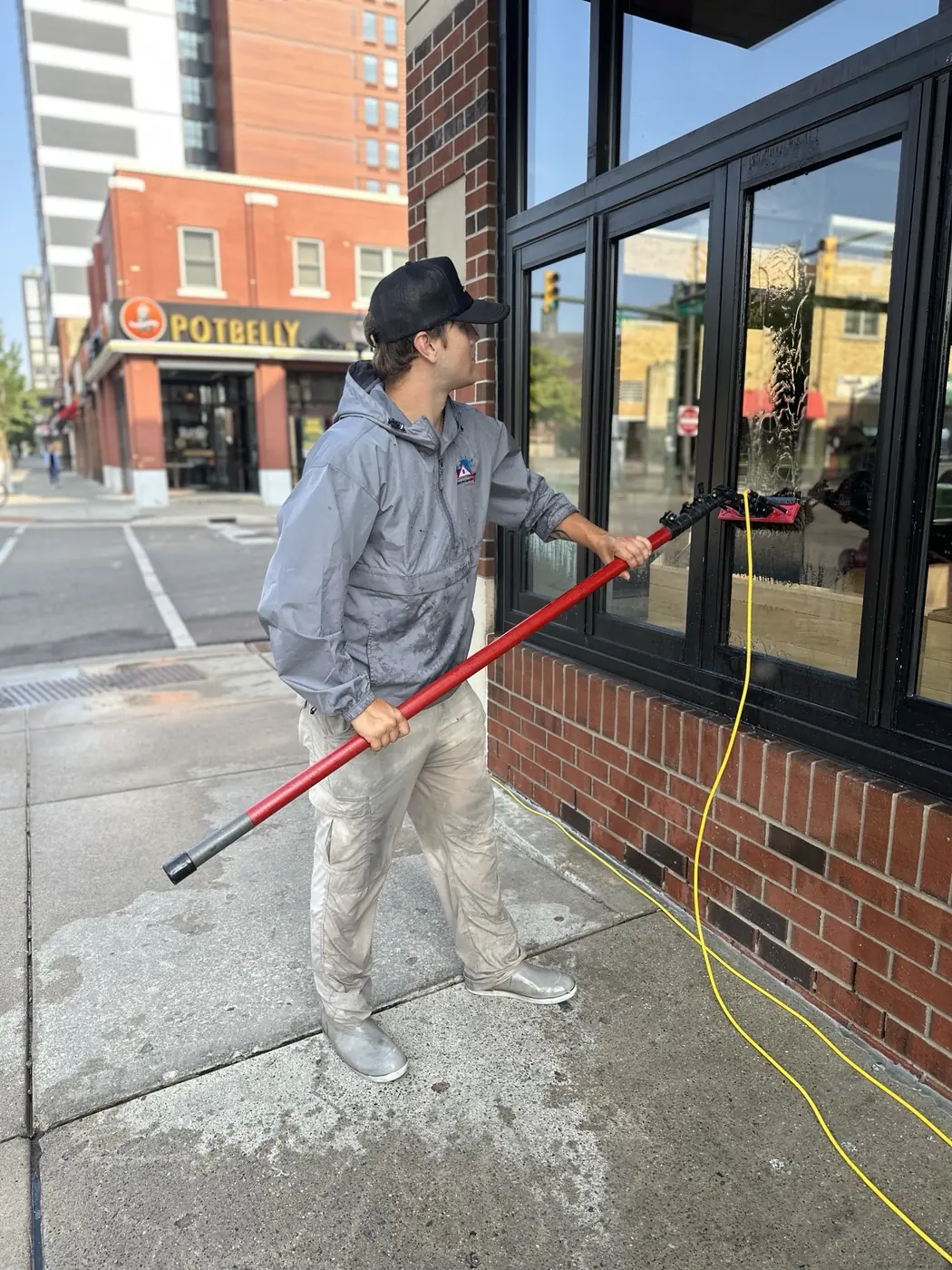 Technician cleaning storefront windows with a water-fed pole and brush from the sidewalk